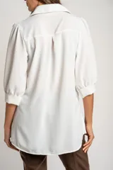 Blusa de corte oversize en color blanco, con cuello camisero, cierre frontal de botones y mangas tres cuartos con sutiles frunces en los hombros.