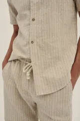 Camisa de manga corta en tejido ligero de lino y algodón a rayas verticales en tonos beige y blanco. Tiene botones de madera y un parche con el logo de la marca.