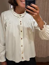 Camisa de algodón color blanco con cuello redondo y detalle de fruncido tipo nido de abeja en el canesú. Presenta cierre frontal con botones decorativos, mangas largas abullonadas con puños elásticos fruncidos y volados en el cuello.