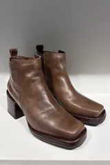 Bota corta de cuero color marrón, con diseño de punta cuadrada y taco cuadrado con textura grabada. Presenta costuras visibles y una lengüeta en la parte posterior.