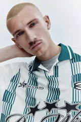 Camiseta de fútbol de manga corta en malla estampada, color blanco con diseño a rayas verticales en tono verde y detalles gráficos en el pecho. Cuenta con cuello abierto verde con finas rayas blancas y corte holgado.