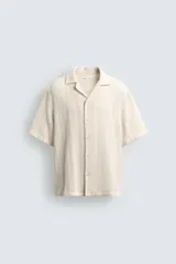 Camisa de calce relajado confeccionada en tejido con algodón de color beige. Presenta cuello bowling, manga corta, aberturas laterales en el bajo y cierre frontal con botones.