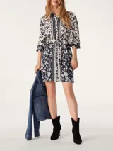Vestido camisero corto de Ba&sh con estampado floral en tonos azul marino y blanco. Presenta cuello camisero, cierre frontal con botones, mangas tres cuartos y un lazo ajustable en la cintura que define la silueta.