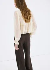 Camisa de diseño recto en tono beige, confeccionada con tejido combinado y panel de encaje frontal. Presenta cuello redondo, mangas largas abullonadas con puños elásticos y detalle de volantes en la cintura.