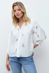 Blusa de color crudo con escote en V y mangas tres cuartos abullonadas. Presenta un delicado diseño de flores bordadas en tonos azules y amarillos distribuidas por toda la prenda.