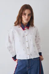 Camisa de lino blanca con estampado de motivos deportivos, cuello y puños tejidos a mano en crochet azul con borde rojo. Presenta cierre frontal con botones y un bolsillo en el pecho.