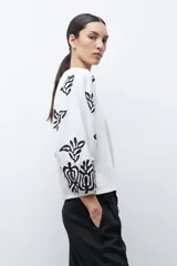 Remera de color crudo con mangas largas abullonadas, decoradas con bordados en color negro. Presenta un diseño de cuello redondo y corte recto.