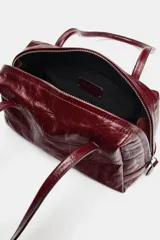 Bolso de hombro de piel con formato bowling, acabado texturizado tipo cocodrilo y color bordó. Cuenta con doble asa de hombro, cierre principal de cremallera con doble tirador y bolsillo interior con cremallera.