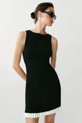 Vestido corto de punto canalé negro, con cuello barco, sin mangas y detalle de volados blancos en el borde inferior.