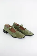 Zapatos tipo Mary Jane de cuero con textura efecto avestruz en color verde oliva. Presentan una tira en T con cierre de hebilla circular al tono, punta cuadrada y un taco bajo cuadrado de color negro.