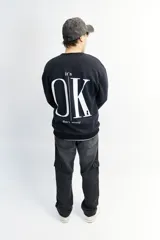 Buzo negro de corte oversize con cuello redondo y puños ajustados. Estampa frontal con la frase "It's OK" en letras blancas.