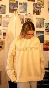 Buzo de tejido de punto color crudo con rayas verticales finas en tono beige. Presenta un diseño holgado con cuello redondo y el texto 'GLASSY WAVES' bordado en el centro del pecho.