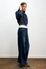 Chaqueta estilo bomber confeccionada en tejido denim de color azul oscuro. Presenta cuello redondo, manga larga, bolsillos delanteros de vivo y cierre frontal con botones ocultos.