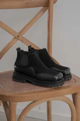 Botas tipo chelsea de caña baja, confeccionadas en una combinación de cuero gamuzado y cuero texturizado en tono marrón oscuro. Presentan una suela track de plataforma gruesa y elásticos laterales negros.