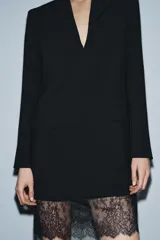 Vestido tipo blazer negro, de corte recto y largo medio, con cuello solapa, manga larga con abertura y botones, y bolsillos con solapa en el delantero. Presenta un bajo con aplicación de encaje negro a tono.