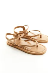 Sandalia plana color marrón claro, estilo gladiadora minimalista, con tiras finas adornadas con tachas doradas. Presenta un diseño de dedo (tira entre los dedos) y tiras que se atan o ajustan alrededor del tobillo con hebilla dorada.