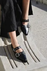 Zapato de cuero negro con punta fina, dos tiras con hebillas plateadas y detalle de ojales metálicos.