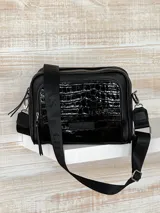 Bandolera negra de cuero con textura tipo cocodrilo en la parte superior. Tiene correa de tela negra con letras blancas y herrajes plateados.