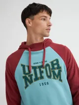 Canguro negro y ocre con capucha y cuello en rib, manga raglán, bolsillos laterales y estampa combinada (estampa y bordado).