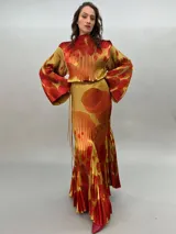 Vestido largo plisado con estampado abstracto en tonos rojo, naranja y dorado, cuello alto y mangas acampanadas. Incluye lazo fino para ajustar en la cintura.