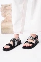 Sandalia negra de plataforma gruesa con dos tiras anchas ajustables con hebilla. Una de las tiras tiene un estampado de vaca blanco y negro.