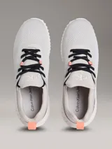 Zapatillas tipo runner Calvin Klein, de color gris con detalles en negro y coral. Presentan un diseño de malla perforada en la parte superior, cordones negros, suela chunky de EVA blanca y el logo de la marca en la lengüeta y el lateral.