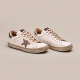 Zapatillas bajas de cuero vacuno blanco con aplique de estrella en cuero color aluminio y talón en cuero plateado. Cordones de algodón encerados color gris y suela de caucho con banda cosida. Plantilla de goma eva de 17 mm.