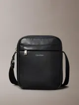 Cartera reporter estructurada Calvin Klein de poliuretano con textura granulada, color negro, cierre superior de cremallera y bolsillo frontal con cierre. Cuenta con correa de hombro ajustable y logo metálico de la marca en la parte delantera.