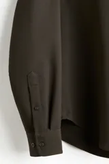 Camisa de manga larga color marrón oscuro, confeccionada en sarga suave, con cuello americano y tapeta clásica de botones. Presenta canesú en la espalda y bajo redondeado.