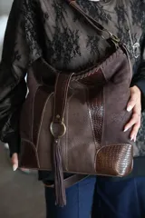 Bolso marrón chocolate confeccionado en cuero vacuno, con diseño de paneles combinados de cuero liso, texturizado y con grabado efecto cocodrilo. Cuenta con manija corta, correa larga ajustable y removible, y detalle de argolla metálica con borla de flecos colgante.