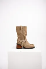 Bota de caña media confeccionada en cuero gamuzado color chocolate. Presenta un diseño de estilo western con taco cuadrado de 7 cm y una correa decorativa en el tobillo con herrajes metálicos plateados.