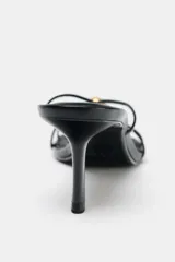 Sandalia de tacón fino color negro, con tiras finas en la parte delantera y detalle de aplique metálico dorado.