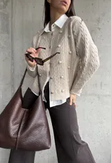 Cardigan de punto color beige con diseño de trenzas y pompones decorativos. Presenta cuello redondo, cierre frontal con botones y mangas largas.