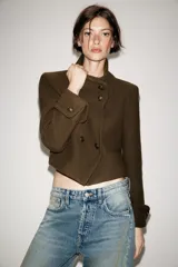 Chaqueta corta de paño color verde militar, con cuello subido, manga larga con puños abotonados y cierre frontal cruzado con botones metálicos. Presenta hombreras y bolsillos de vivo en el delantero.