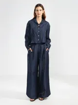 Camisa de manga larga color azul marino, confeccionada en lino italiano con hilo metalizado que le da un sutil brillo. Presenta cuello clásico y botones frontales, con un detalle de broche o dije en forma de flor en el cuello.