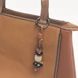 Cartera tipo tote color camel, con paneles laterales de textura granulada y panel central de tejido con textura diagonal. Incluye doble asa de mano, correa larga ajustable y extraíble, cierre superior y un dije colgante decorativo con cuentas de madera y piedra.