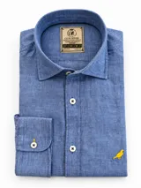 Camisa de lino color azul, manga larga, con cuello corto clásico, botones blancos con costuras en cruz y bordado de un ave amarilla en el pecho.