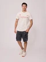 Remera color crema de hombre, marca Rusty, con estampado frontal en color rojo con el logo de la marca y la leyenda "Rusty Worldwide Supply Co. Since 1985".