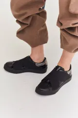 Zapatillas urbanas de color negro con diseño slip-on, caracterizadas por bandas elásticas cruzadas en el empeine y detalles en charol. Cuentan con suela plana de goma al tono.
