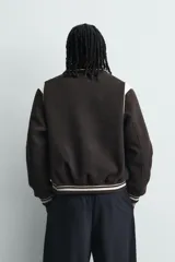 Cazadora estilo varsity de corte cropped, confeccionada en tejido de lana marrón con mangas en contraste color crema. Presenta cuello solapa, cierre frontal con botones a presión, bolsillos laterales de vivo y acabados en rib a rayas en puños y cintura.