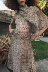 Blusa de cuello subido y manga sisa, confeccionada en tejido fluido semitransparente con estampado de lunares en tonos beige y negro. Presenta un diseño superpuesto tipo capa con caída asimétrica y cierre posterior con abertura y botón.