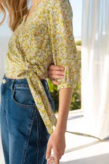 Blusa cruzada con estampado floral en tonos de lila, amarillo y verde. El cruce se ajusta a la cintura con un lazo. Tiene mangas 3/4 con puños elásticos y escote en V.