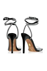 Sandalias de tacón alto color negro con tira delantera transparente de vinilo y tiras plateadas con strass que se atan al tobillo.