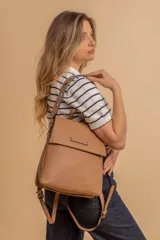 Mochila tipo cartera con tapa, confeccionada en símil cuero color beige. Presenta un diseño moderno con detalle de ojales metálicos en la parte frontal, compartimentos internos organizadores y bolsillo trasero con cierre.