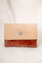 Billetera de diseño bicolor con solapa en cuero gamuzado color beige y cuerpo en cuero liso color marrón, con detalle de aplique metálico circular con logo grabado en el centro.