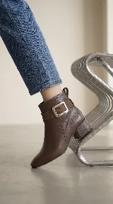 Bota corta de cuero con taco cuadrado bajo y punta redondeada. Presenta una tira decorativa en el tobillo con una hebilla metálica rectangular. Cuenta con cierre lateral interno para facilitar el calce.