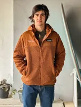 Campera de corderito color naranja con capucha y cierre frontal completo. Cuenta con recortes de tela lisa en el pecho, bolsillos laterales y parches con la marca "La Ola".