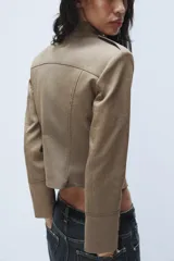 Blazer corto de estilo militar color tostado, con cuello redondo, manga larga y cierre frontal con ganchos metálicos. Presenta detalle de botones decorativos dorados en el frente y en los hombros, con costuras marcadas y forro interior a contraste.