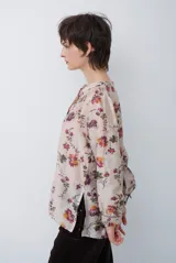 Camisa de corte holgado con estampado floral en tonos rojizos y verdes sobre base beige. Presenta cuello mao con escote en V, cierre frontal parcial con botones y mangas largas con puños ajustados.
