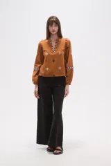 Blusa de algodón color tostado con cuello mao y escote en V. Presenta mangas largas abullonadas con puños elásticos y un diseño con bordados geométricos en contraste distribuidos por toda la prenda.
